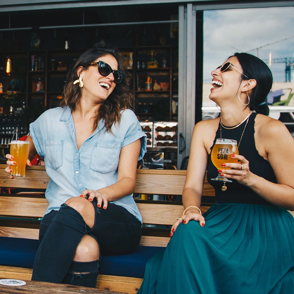 Zwei Frauen mit Sonnenbrillen lachen und trinken Bier auf einer Holzbank in einem Café.