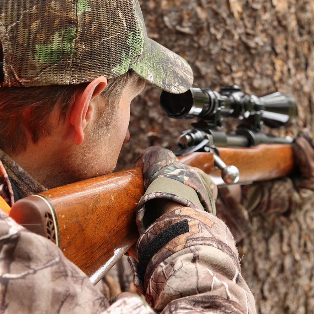 Jäger in Tarnkleidung und -mütze zielt mit einem Gewehr mit Zielfernrohr nahe an einem Baum.