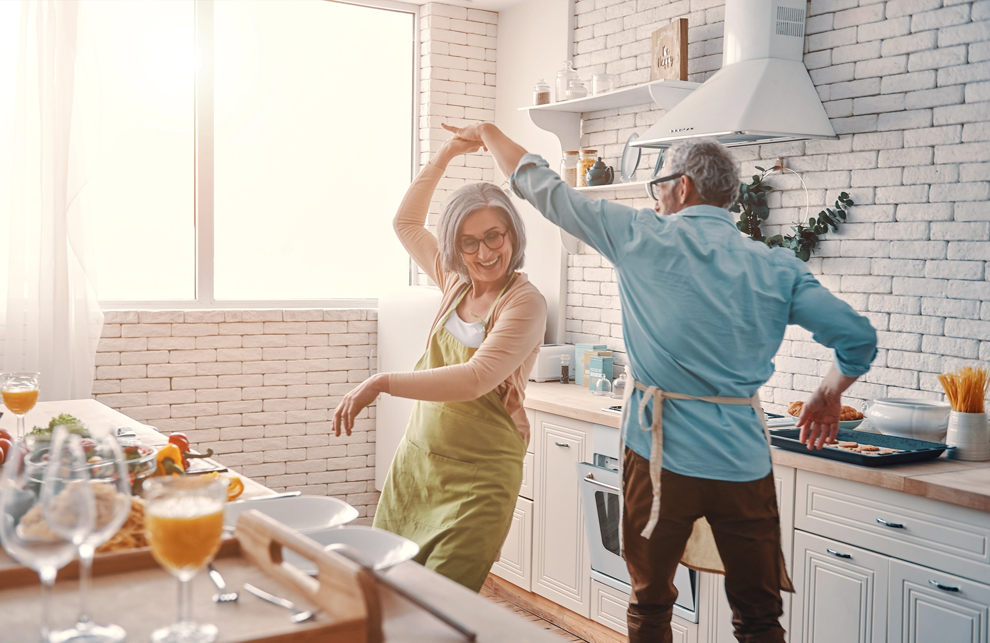 Älteres Paar tanzt fröhlich zusammen in der hellen, modernen Küche beim Kochen.
