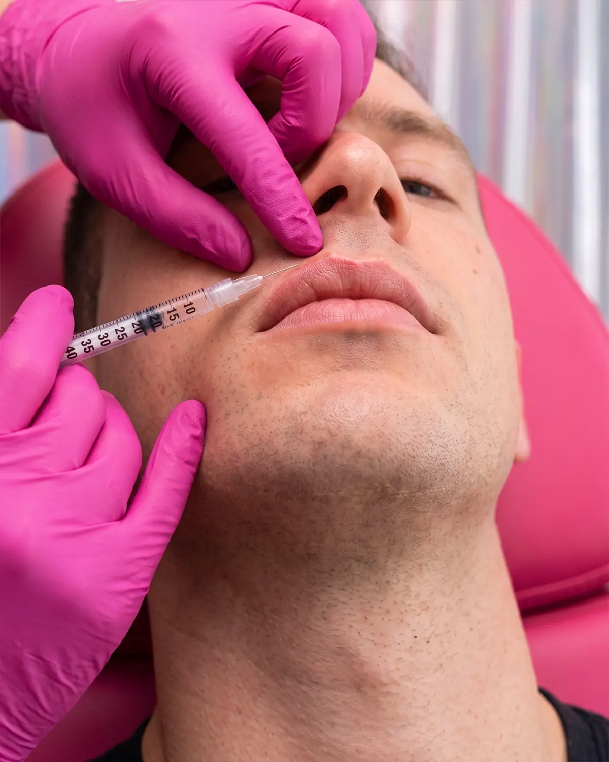 Primo piano di un uomo sdraiato mentre riceve un'iniezione con siringa sulle labbra da mani con guanti rosa.