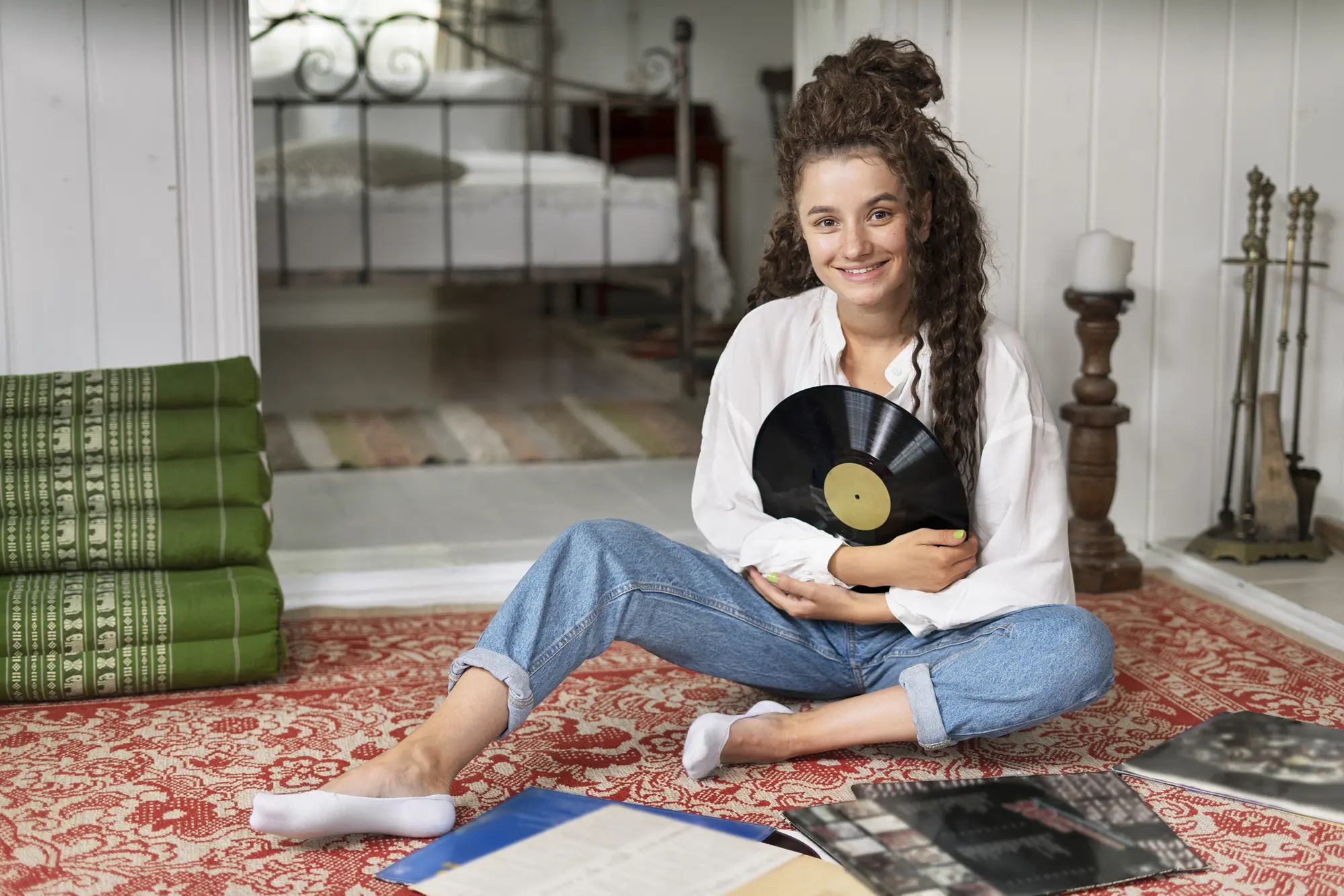 Young woman sitting on a red patterned rug holding a vinyl record and smiling indoors.
