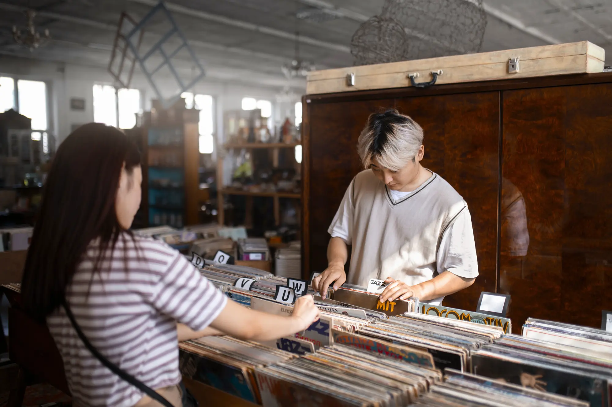 Two people browsing vinyl records in a vintage record store with labeled categories.
