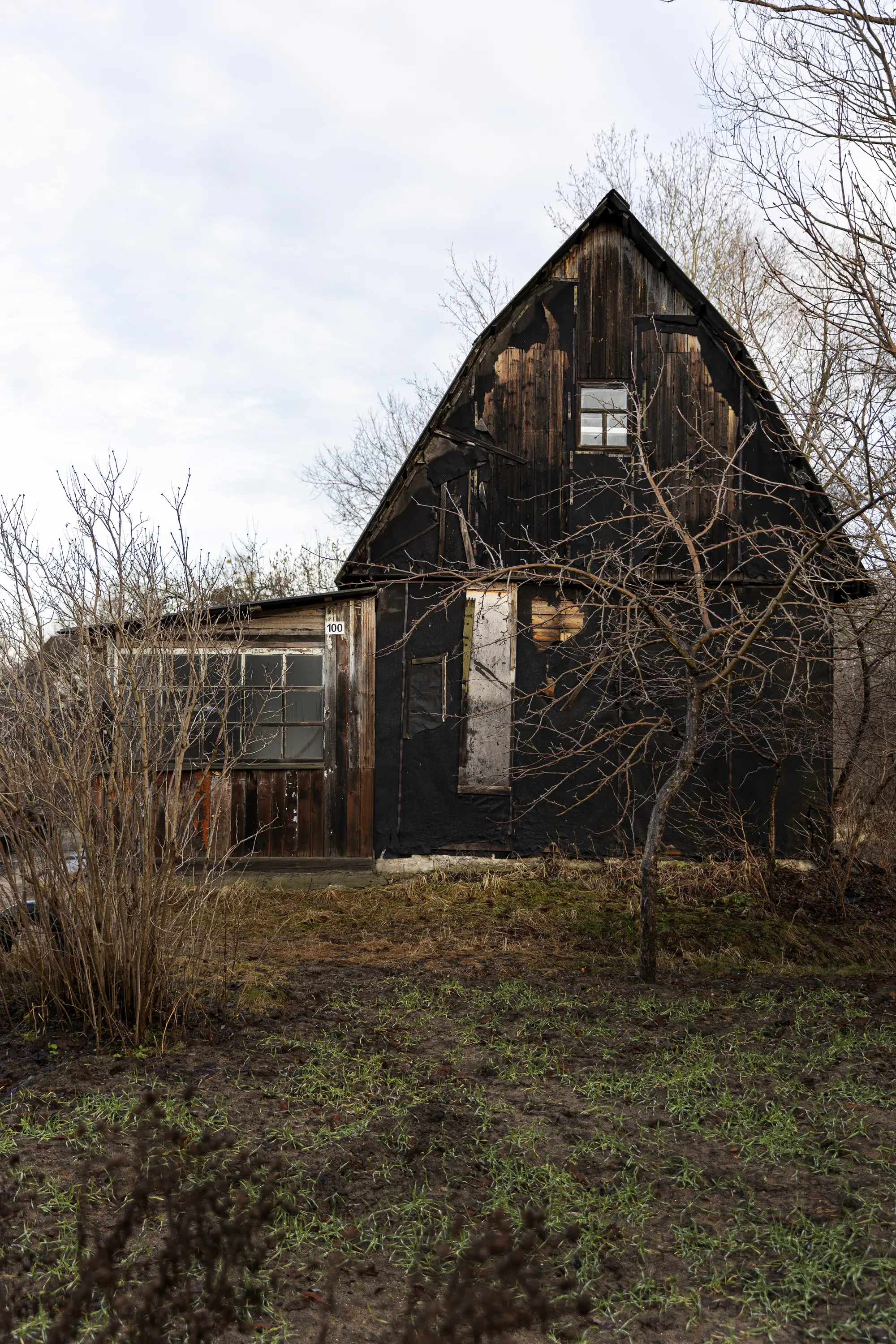 Old wooden house with boarded-up window and leafless trees in front under cloudy sky.