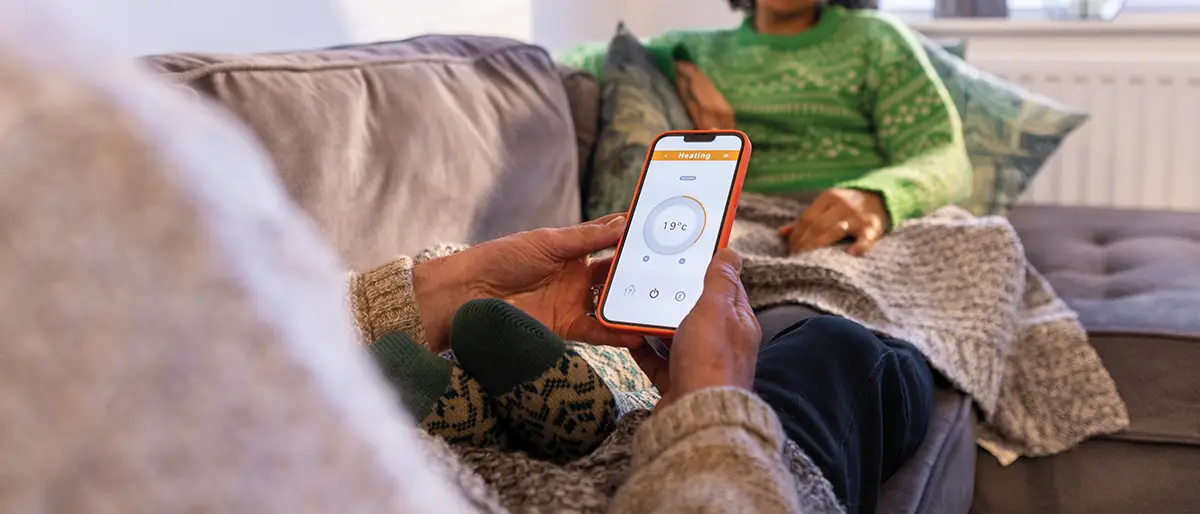 Close-up of hands holding a smartphone set to 19°C on a heating control app on a couch.