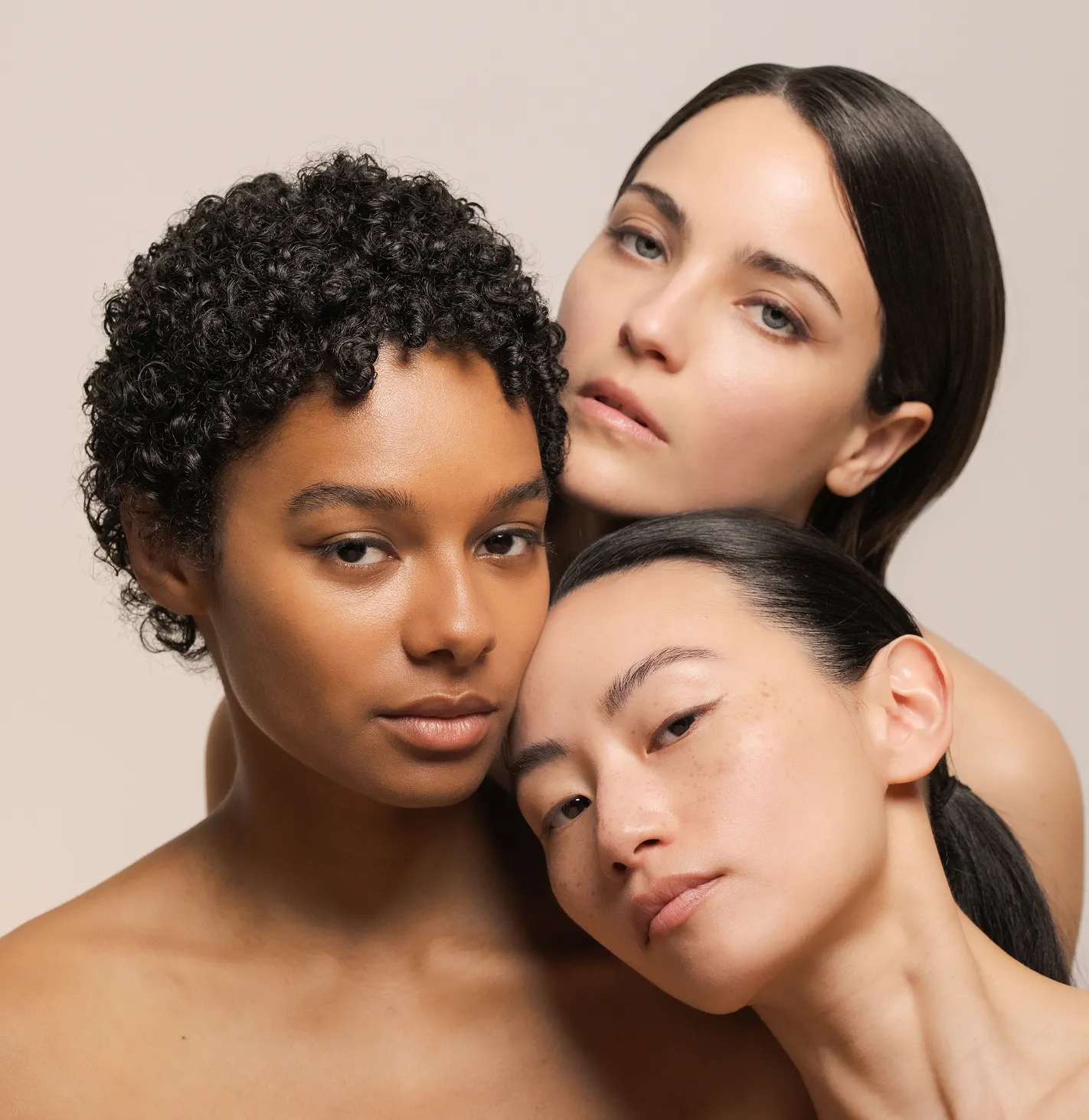 Portrait of three diverse women with eyeliner on, close together with neutral expressions on beige background.