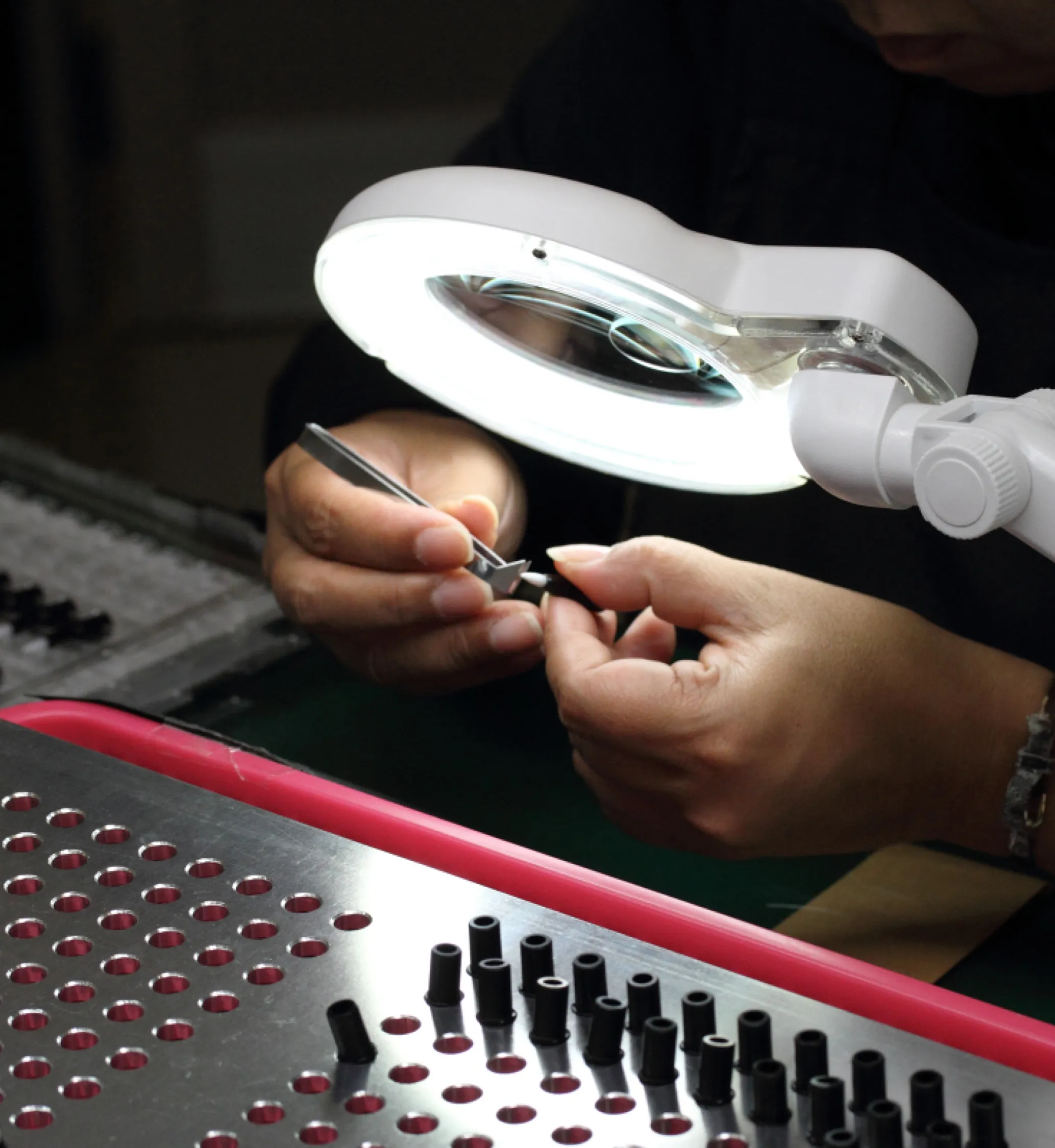 Person inspecting small black components under a magnifying lamp with tweezers.