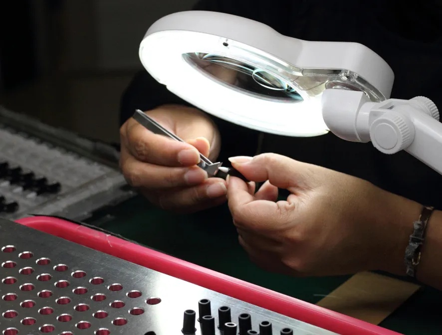 Hands using tweezers to craft or examine a small object under a round magnifying lamp.