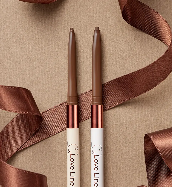 Two brown eyeliner Love liner creamfit pencils with white and beige barrels on a brown surface with ribbon.