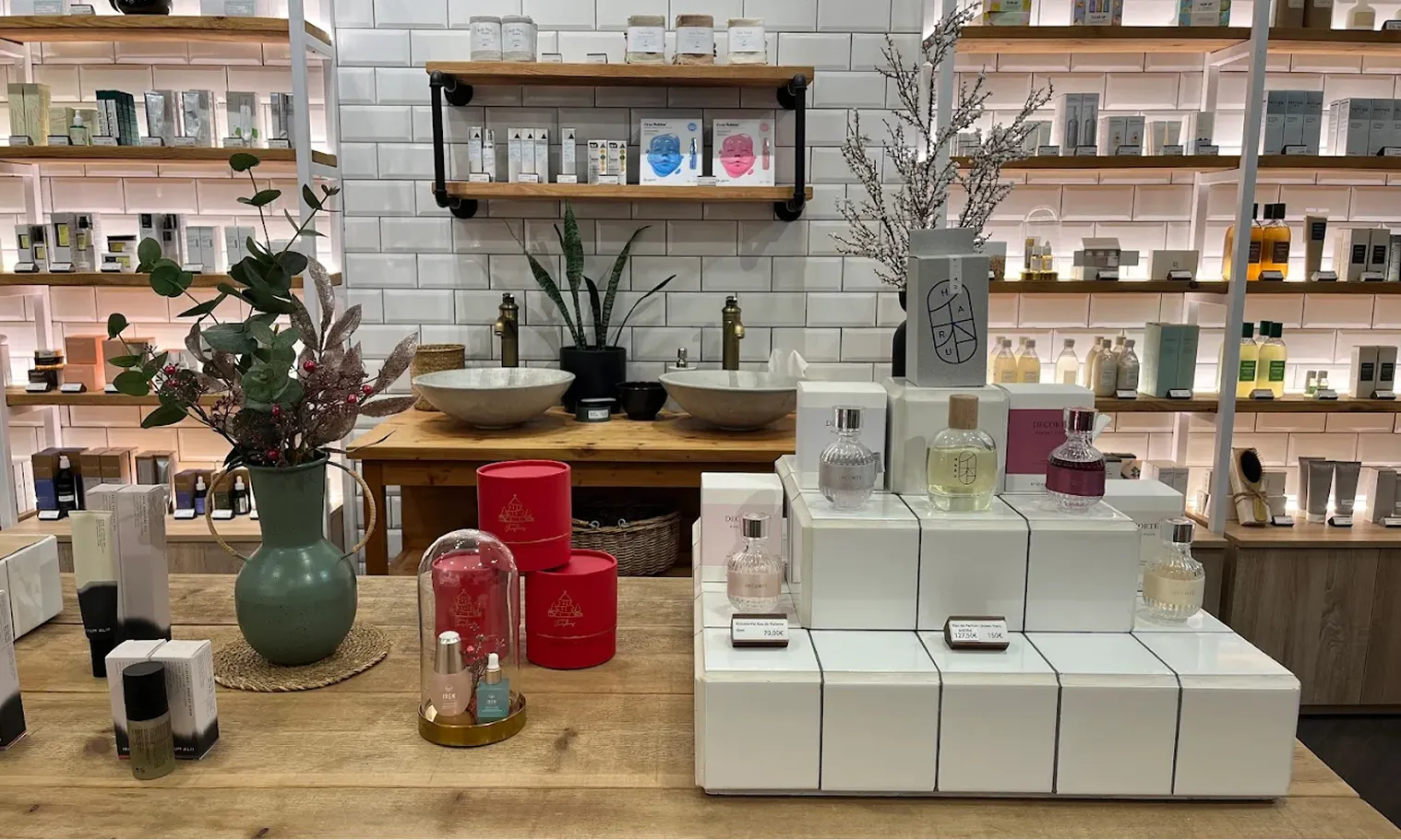Display of perfumes and skincare products on wooden tables with a green vase and two sinks in background.