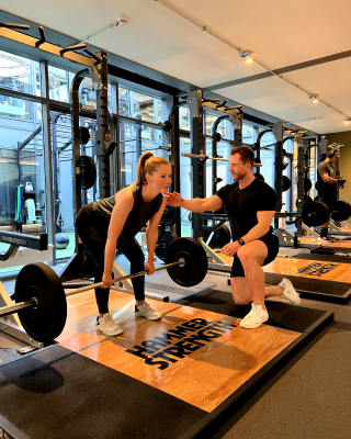 Woman performing deadlift with barbell while trainer kneels and guides in a gym.