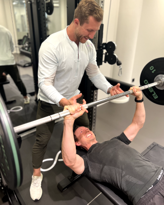 Man lifting barbell on bench press with trainer spotting him in a gym setting.