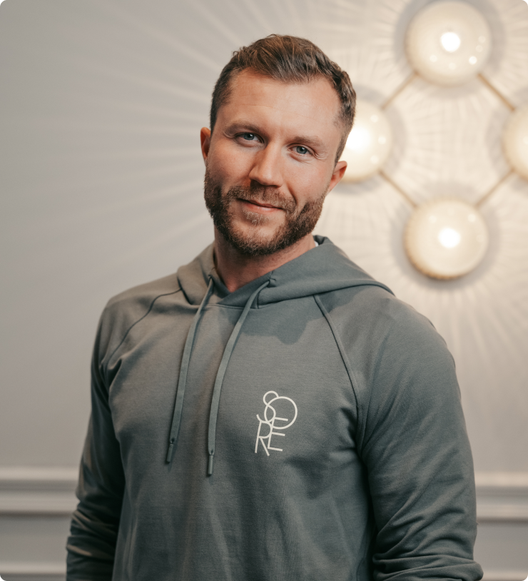 Smiling man with beard in grey hoodie standing indoors near round light fixtures on wall.