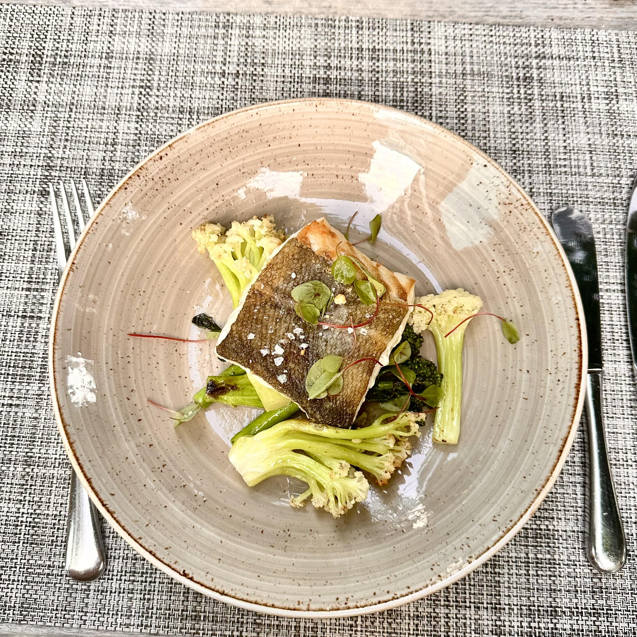 Plated cooked fish fillet with skin, served with grilled cauliflower and greens on a ceramic plate.