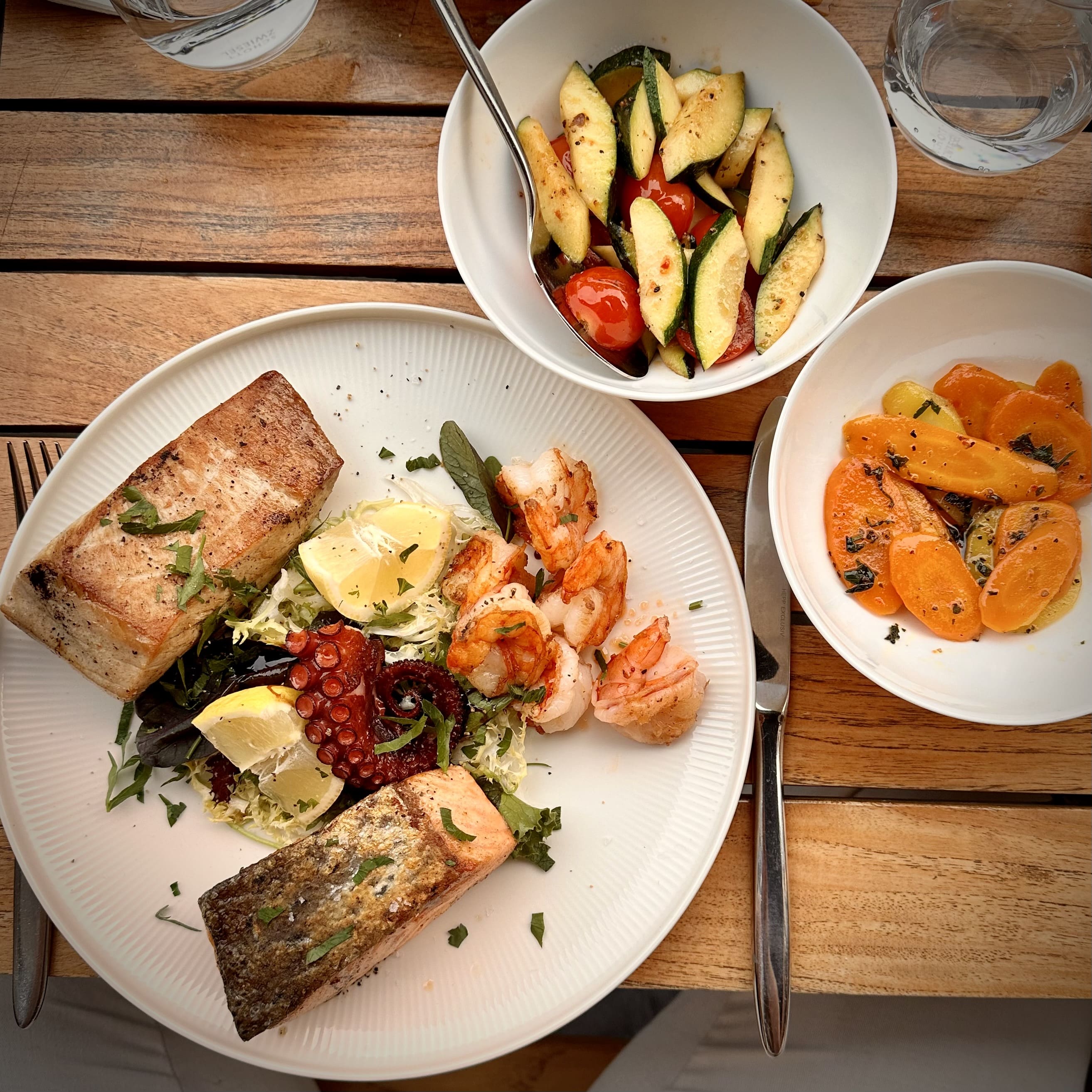 Plate with grilled fish, shrimp, octopus, lemon, and salad; bowls of zucchini tomato and carrots on wood table.