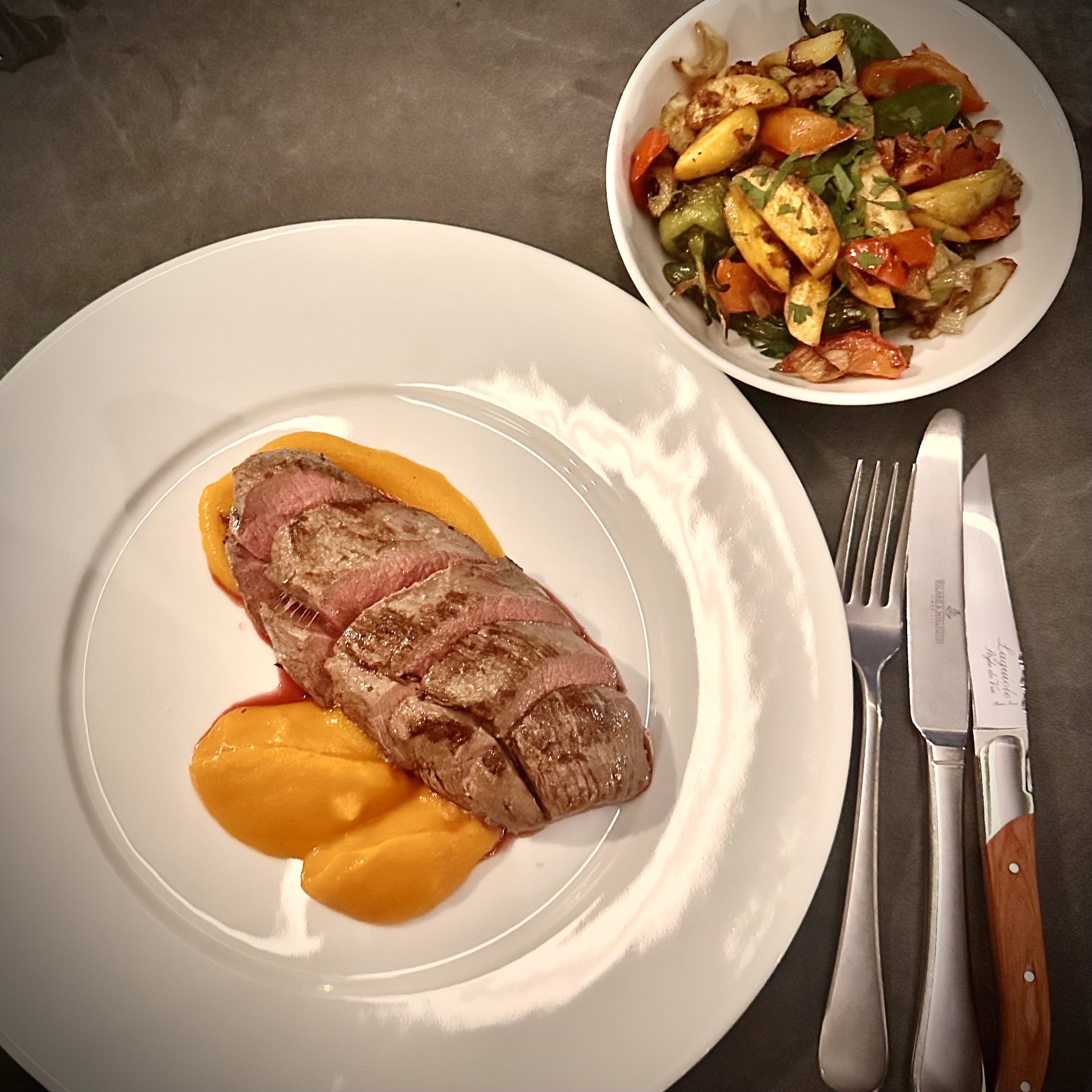 Sliced cooked steak on orange puree with roasted vegetables in a bowl and cutlery on the side.