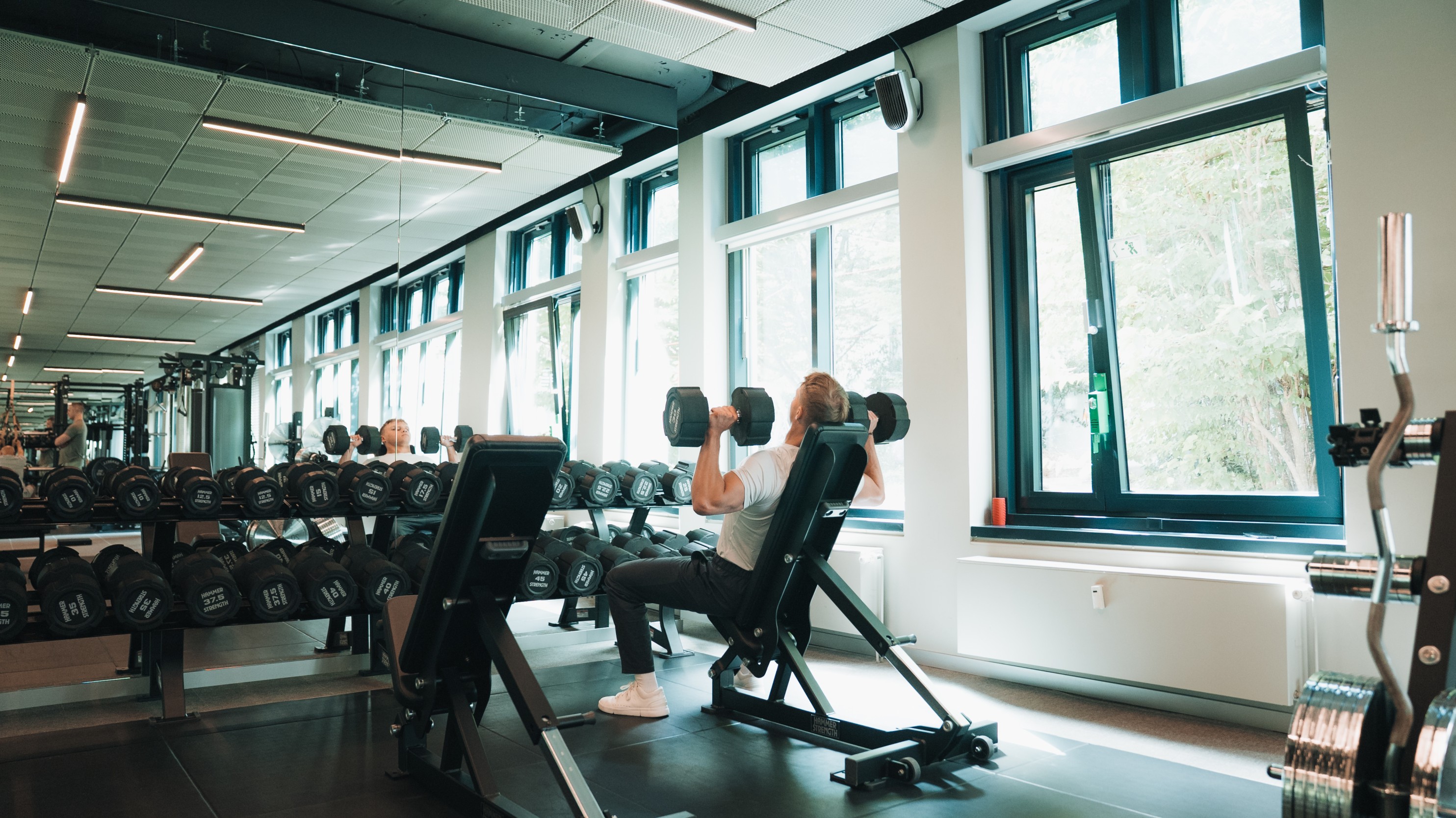 Man doing seated dumbbell shoulder presses in a bright gym with windows and mirrors.