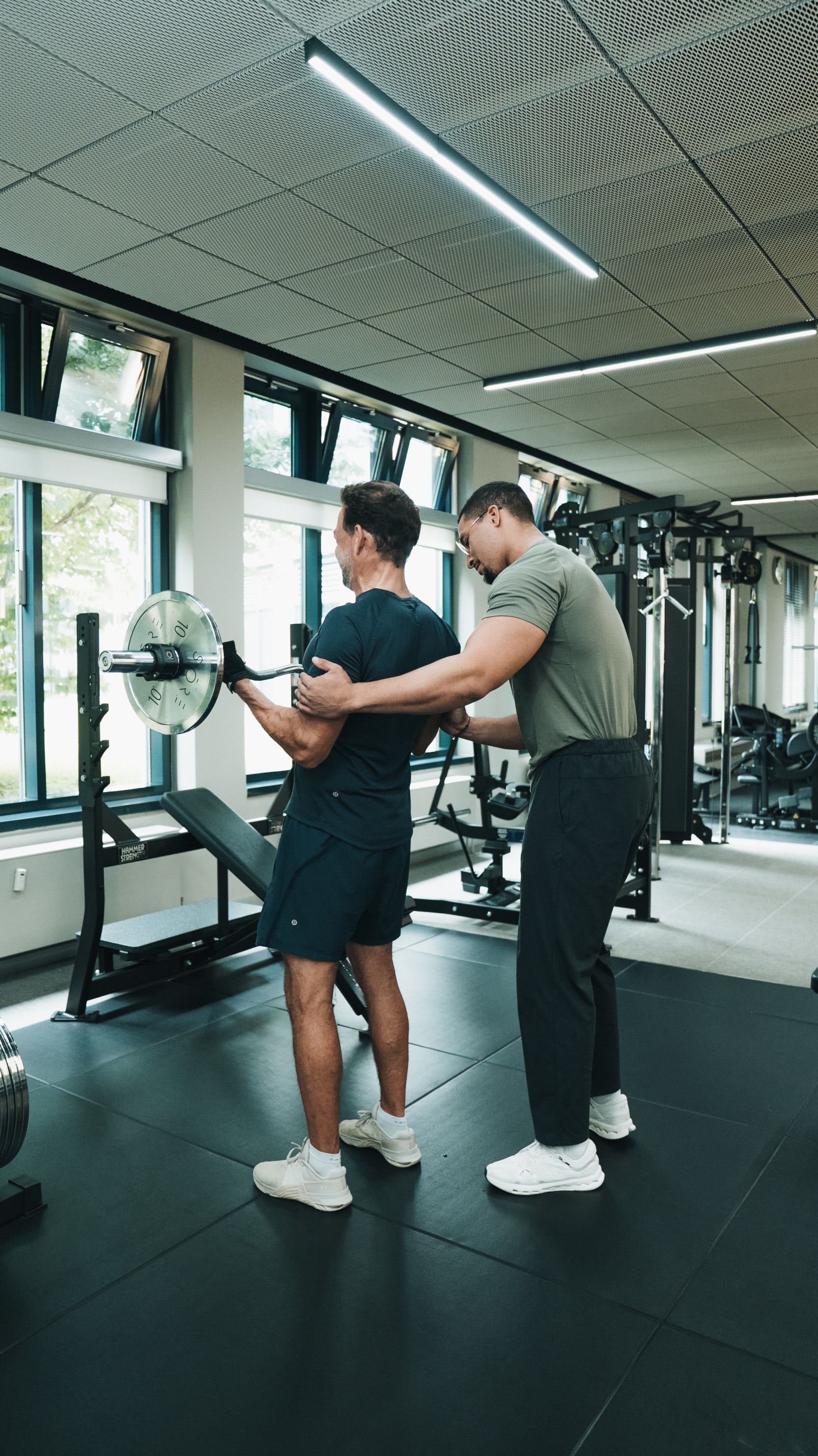 Trainer assists man lifting barbell in gym with large windows and workout equipment.