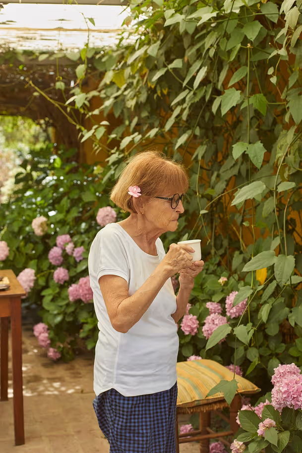 Old lady looking at flowers and drinking tea standing up.