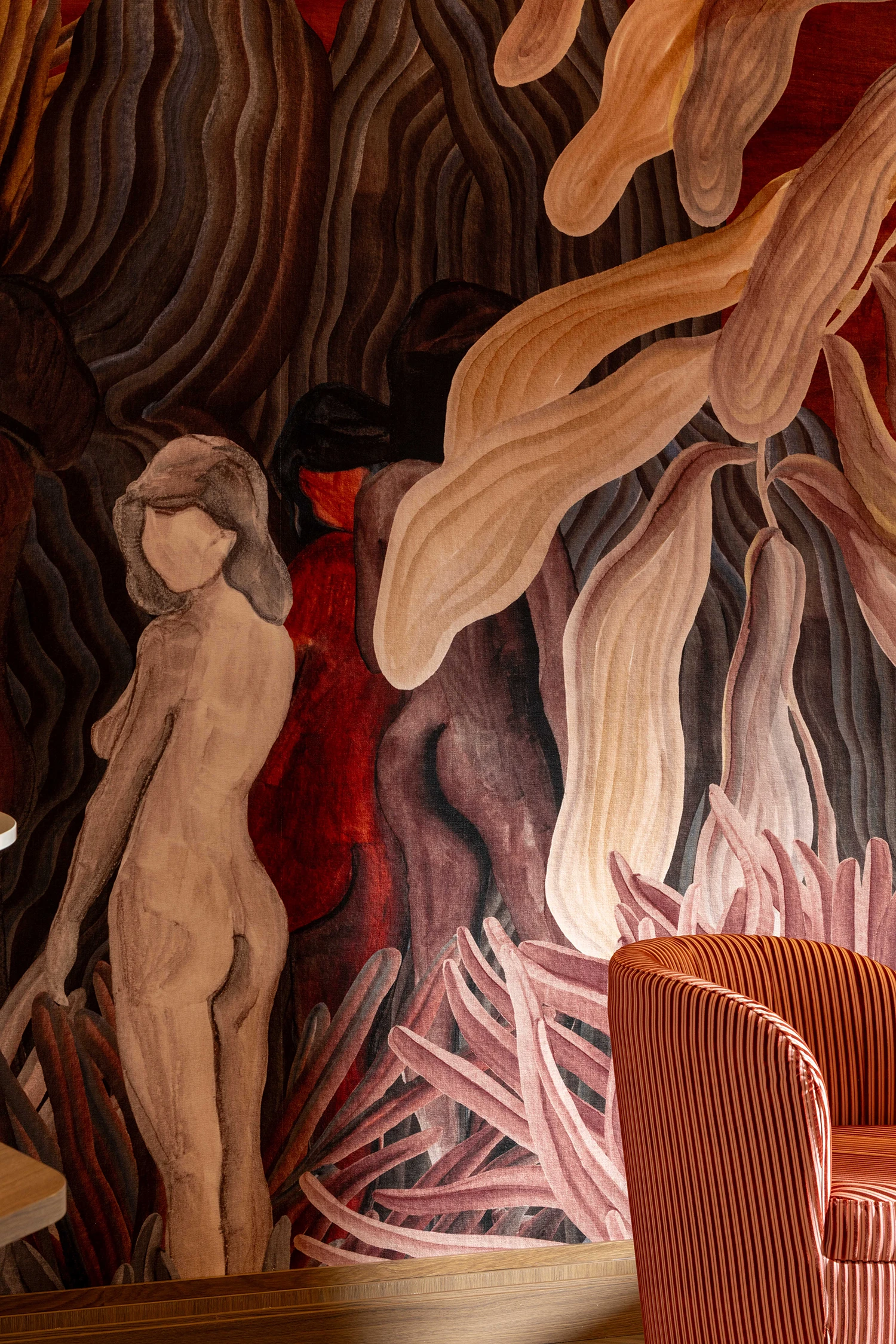 Peinture murale artistique représentant trois figures féminines nues stylisées dans des tons de marron, rouge et beige, entourées de formes organiques ondulantes, à côté d'un fauteuil rayé orange.