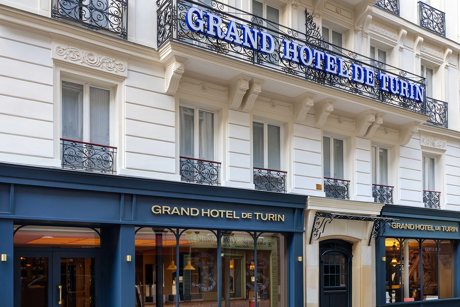 Façade du Grand Hôtel de Turin avec des balcons en fer forgé ornés, des fenêtres à volets blancs, et des enseignes bleues et dorées indiquant le nom de l'hôtel au-dessus et au niveau de la porte d'entrée.