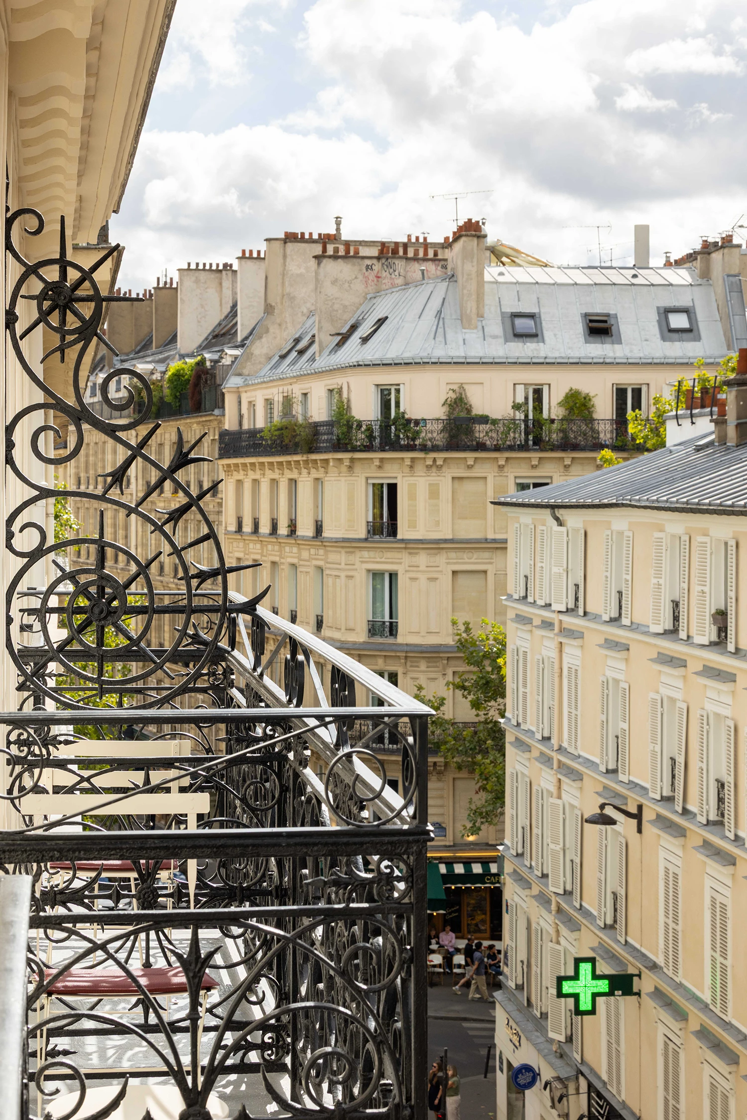 Vue depuis un balcon en fer forgé décoratif sur une rue parisienne avec des bâtiments haussmanniens, des fenêtres à volets, une enseigne de pharmacie verte en forme de croix et un café avec une terrasse en dessous, sous un ciel partiellement nuageux.