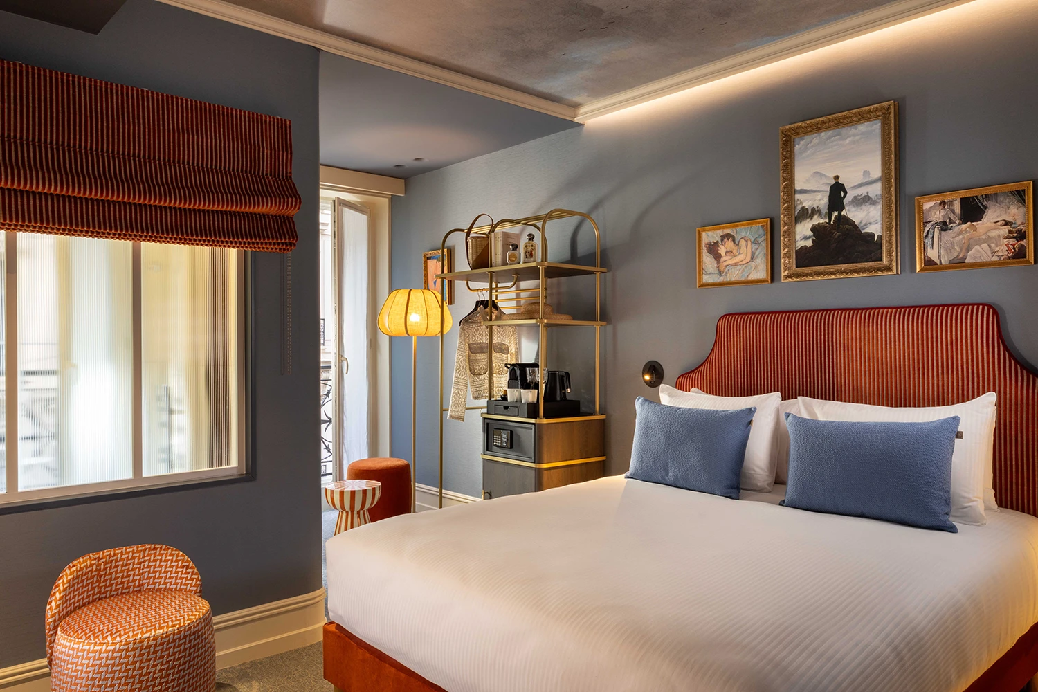 Chambre d'hôtel élégante avec un grand lit à tête de lit rouge rayée, deux coussins bleus, murs gris avec trois tableaux encadrés, lampe sur pied à abat-jour jaune, étagère métallique dorée avec objets décoratifs et fauteuil orange près de la fenêtre.