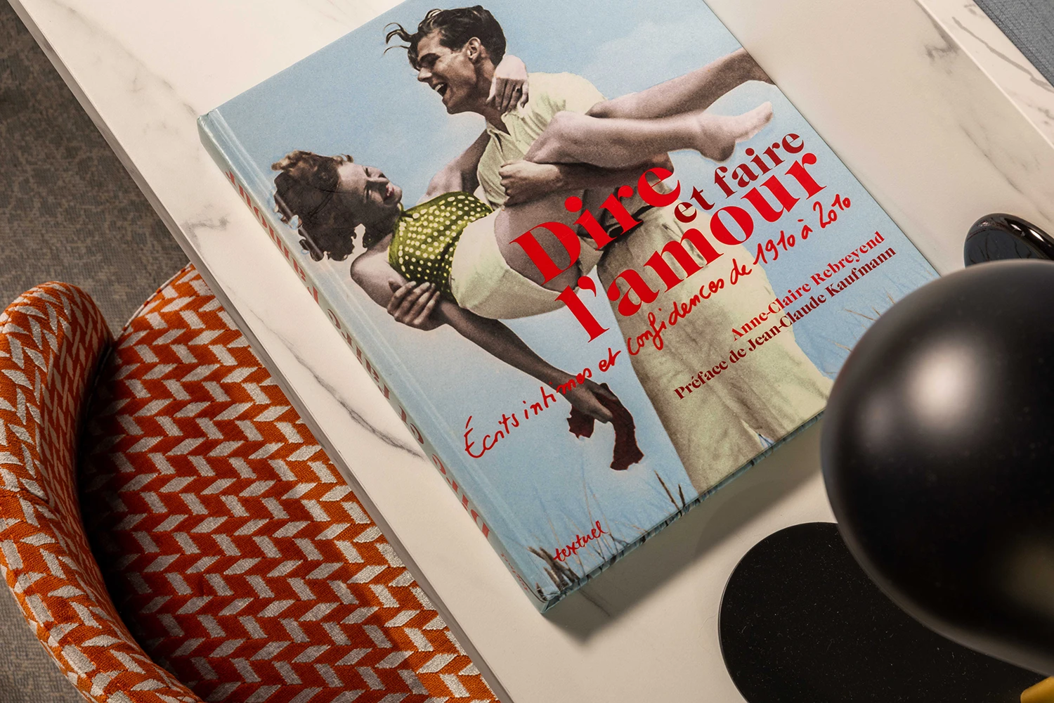 Coin salon avec canapé gris, table basse noire avec livres, fauteuil orange et cheminée noire encadrée d'un miroir doré.