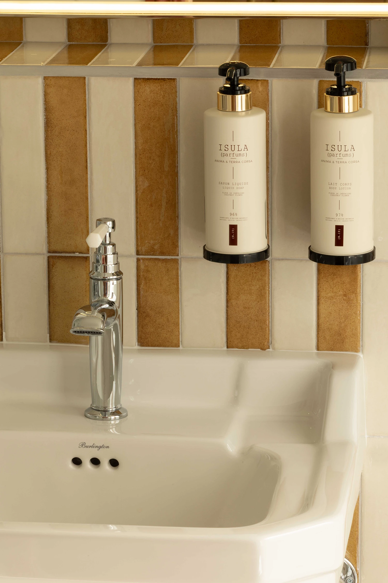 Un lavabo blanc avec un robinet en métal chromé devant un mur carrelé aux tons beige et marron, avec deux distributeurs montés au mur : un savon liquide et un lait corporel marqués Isula parfums.