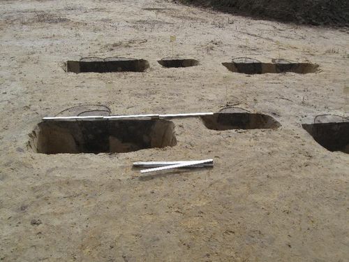 Archeologische opgraving met meerdere rechthoekige greppels en meetlatten in zandgrond.