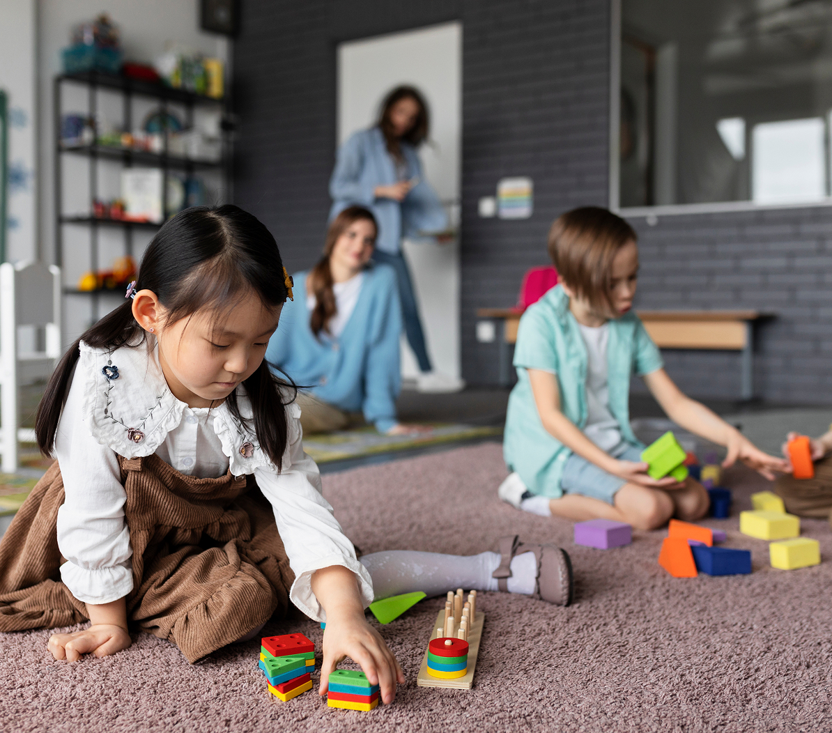 Two children sitting on a carpet in a classroom playing with colorful stacking and building toys, with two adults in the background.