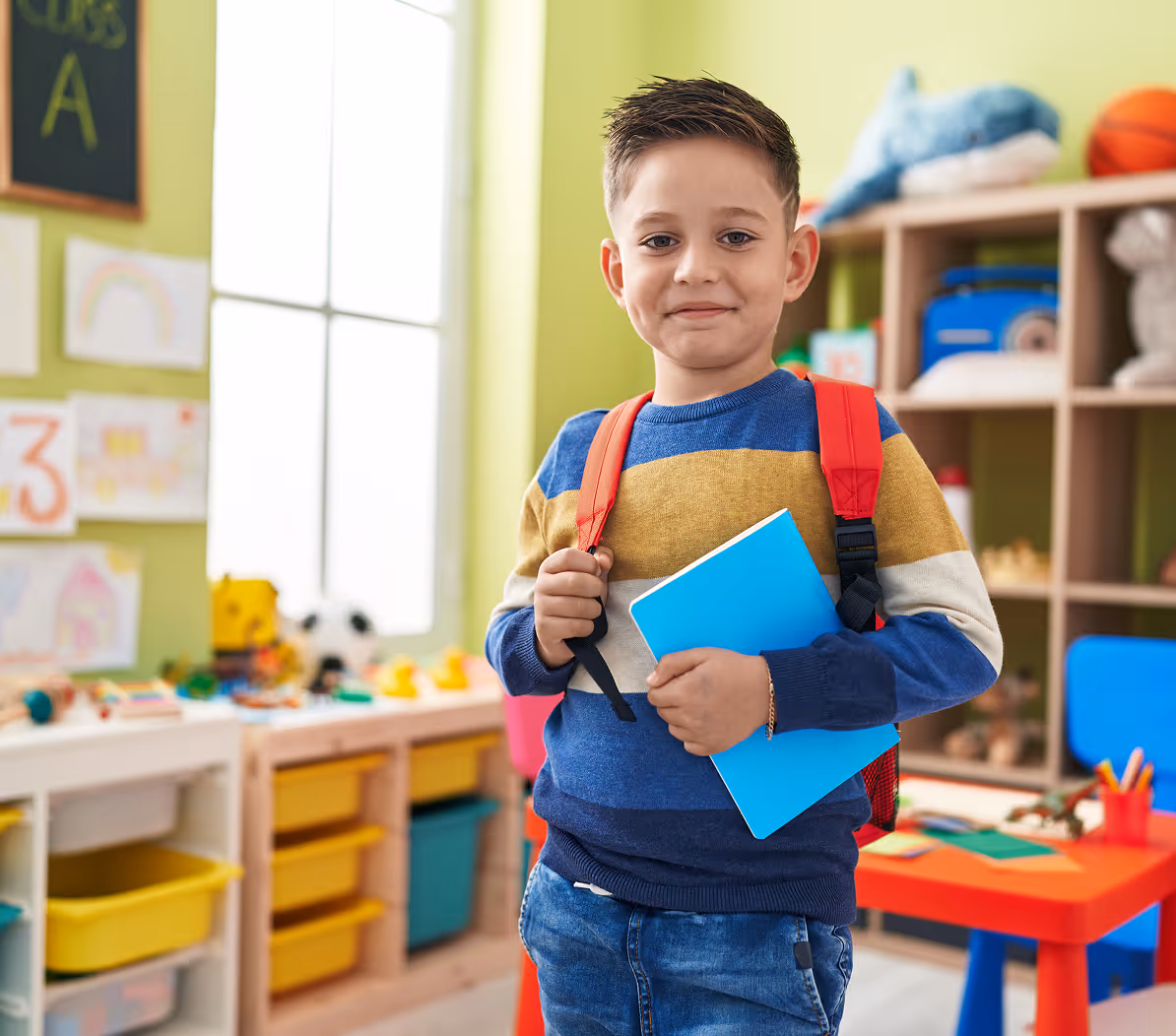 Young boy in a colorful sweater with a red backpack holding a blue notebook in a bright classroom.