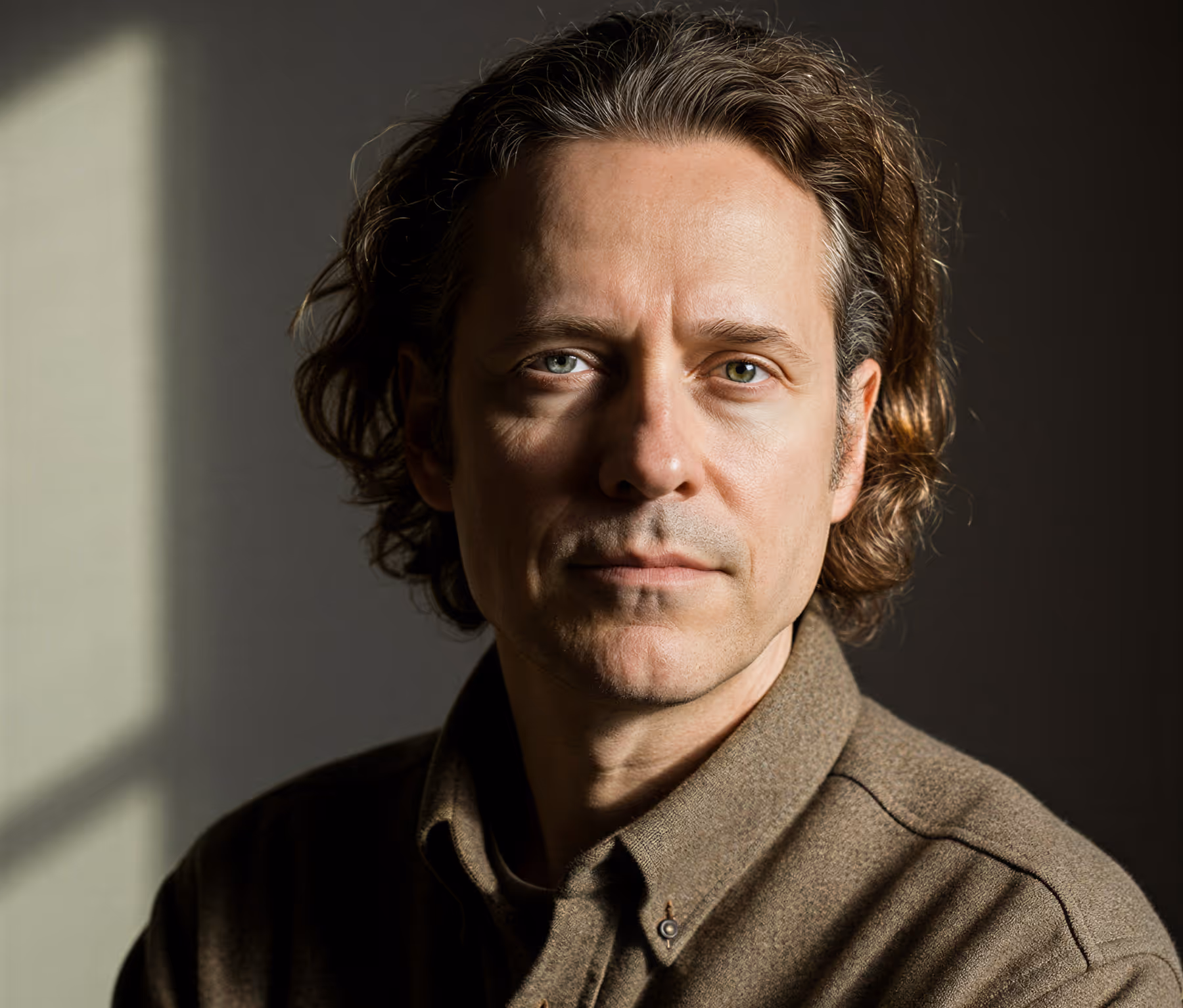 Portrait of a man with curly brown hair wearing a brown button-up shirt, looking thoughtfully at the camera with soft lighting.