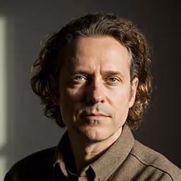 Portrait of a middle-aged man with wavy brown hair, wearing a brown jacket against a dark background.