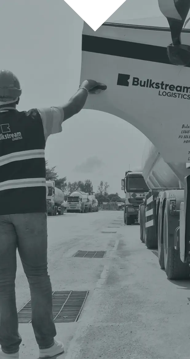 Worker in safety helmet and vest signaling a Bulkstream Logistics tanker truck on a road with trucks.