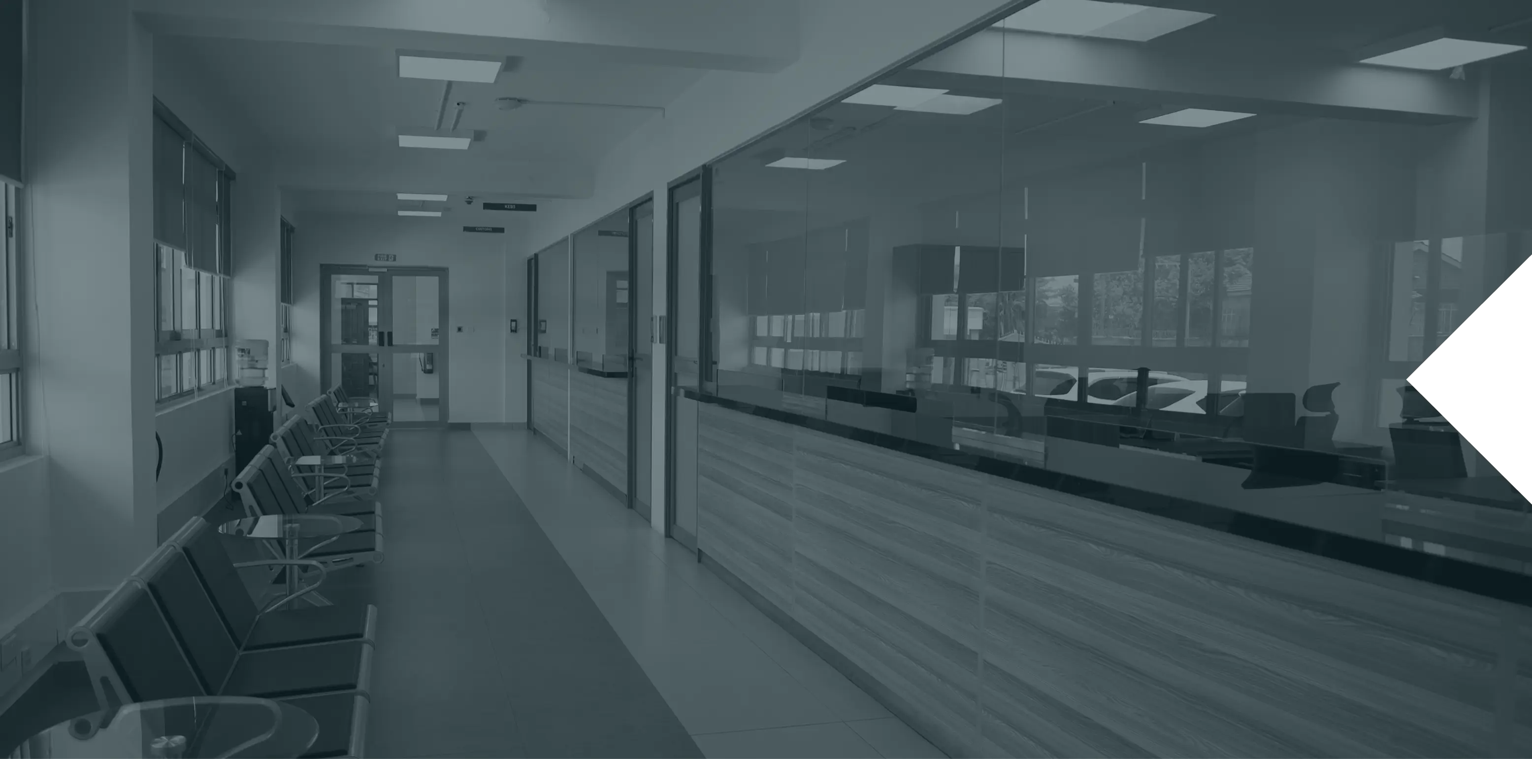 Empty hallway in a modern office or clinic with rows of black chairs on the left side and glass windows with wooden panels on the right, leading to a door at the far end.