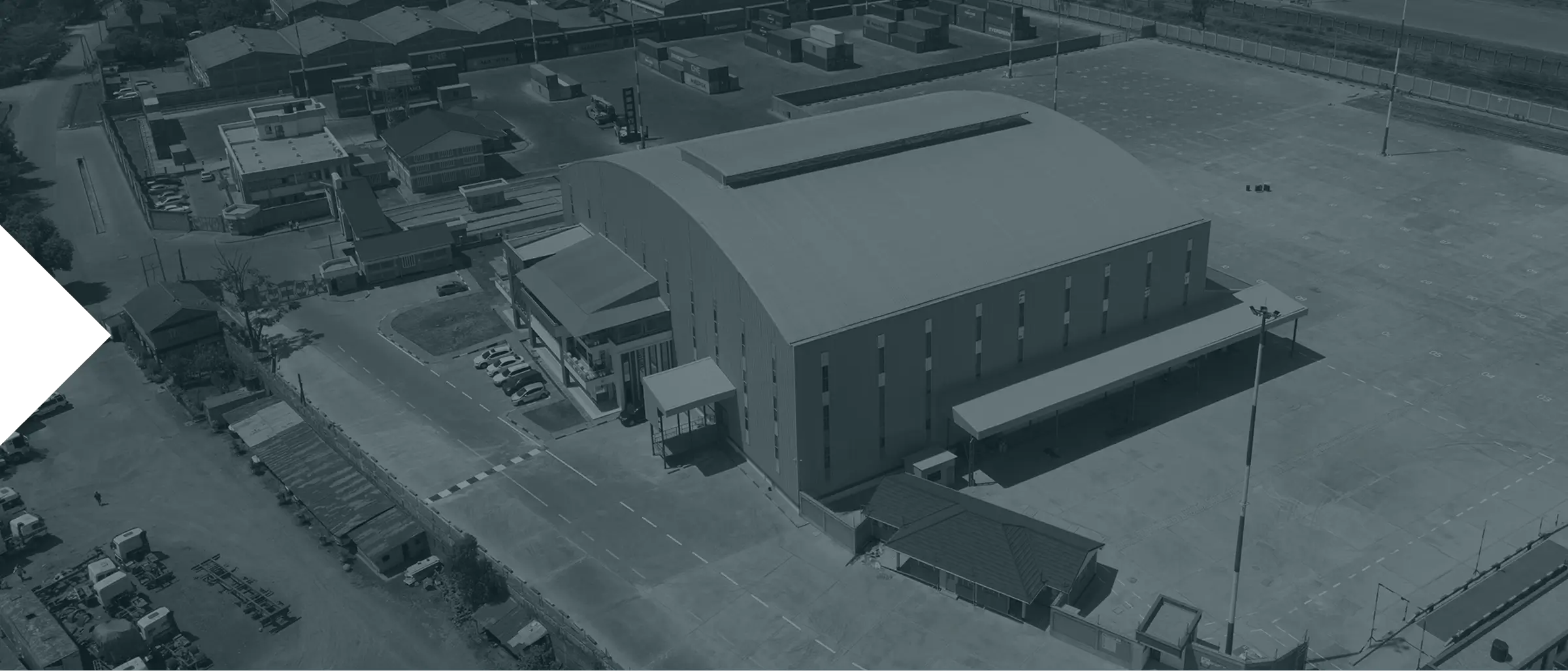 Aerial view of a large industrial facility featuring a wide arched-roof warehouse with multiple windows and adjoining office buildings surrounded by a spacious paved yard and parked vehicles.