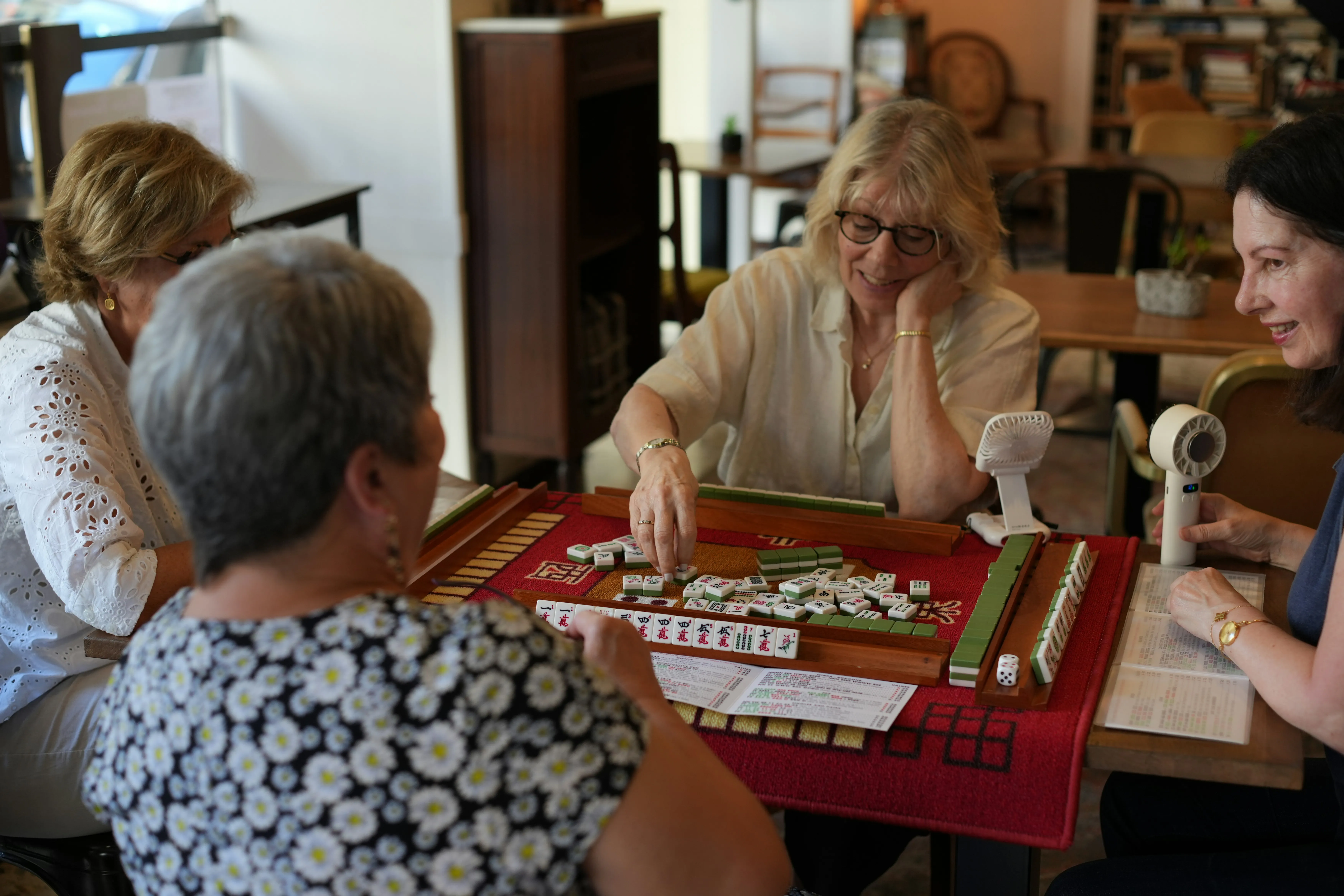 Quatre femmes jouent au mahjong autour d'une table rouge dans une pièce chaleureuse.