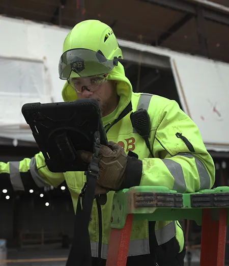 A construction worker in neon safety gear uses a tablet while standing on a ladder.