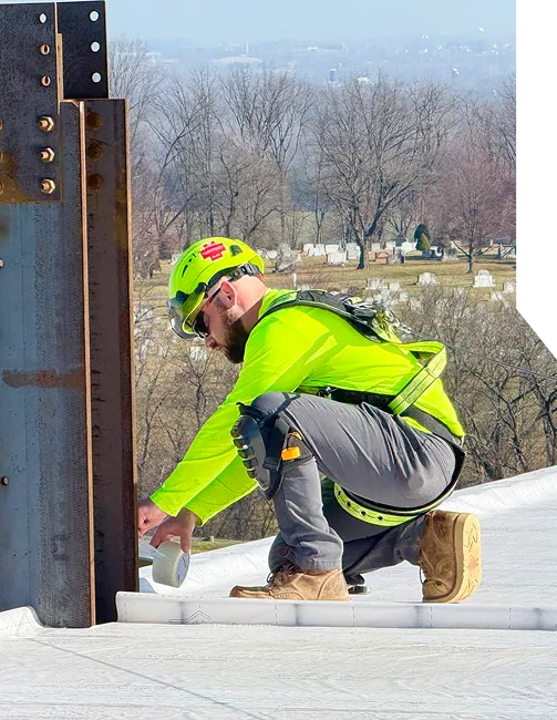 A construction worker in safety gear applies tape to a rooftop edge.