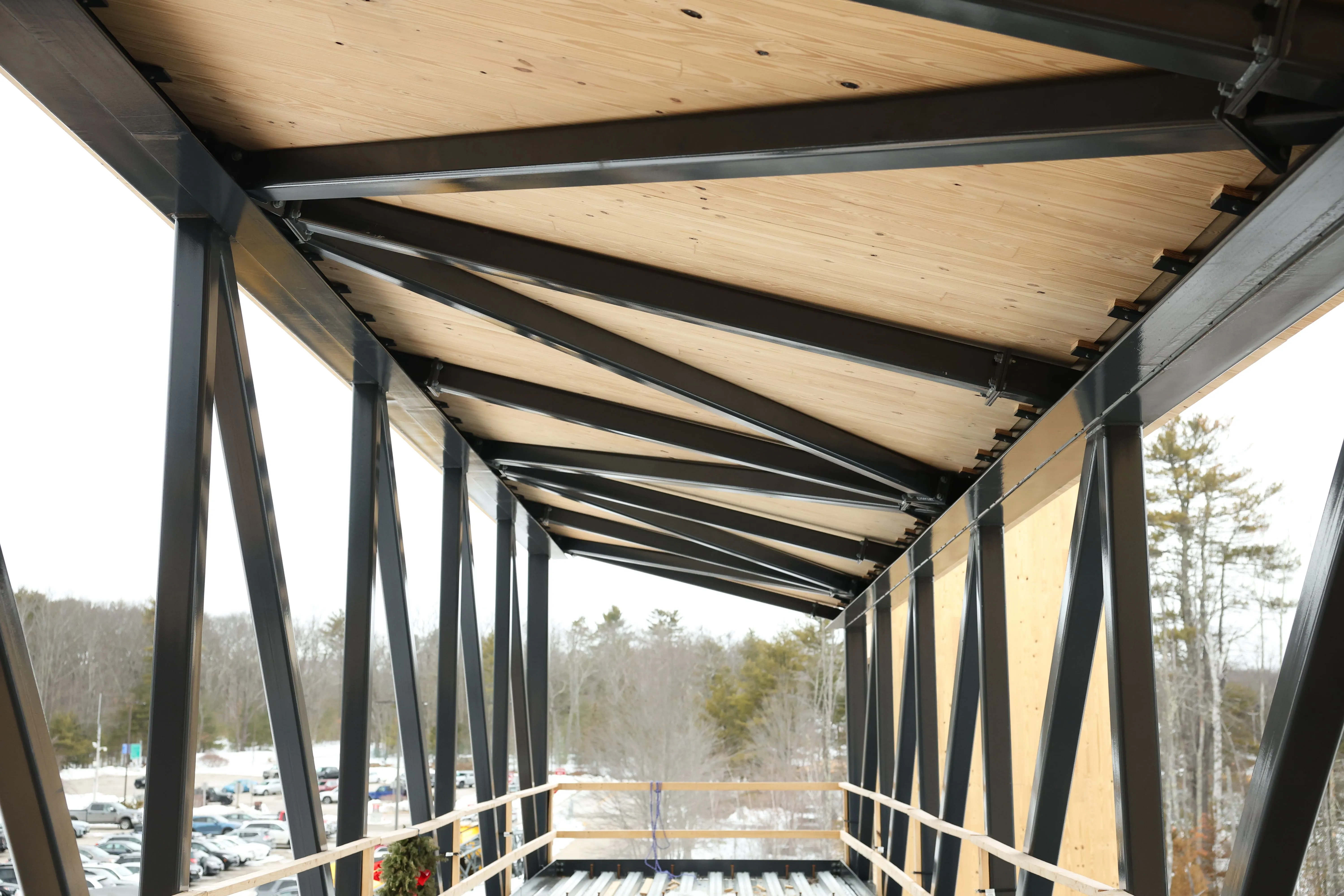 View along the ceiling and black metal beams of a covered outdoor walkway overlooking parked cars and trees.