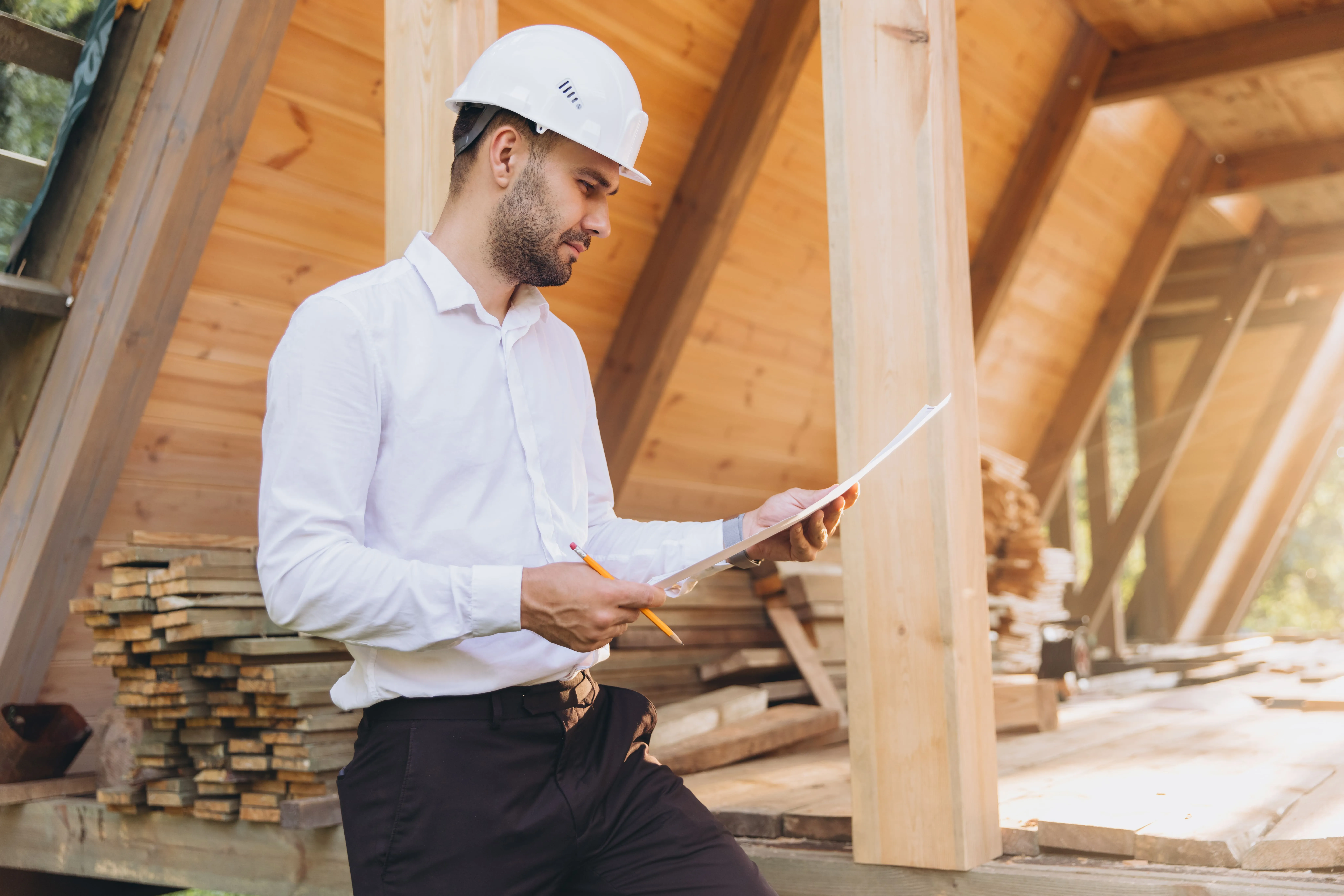 Construction worker in a white helmet reviews blueprints at a wooden building site.