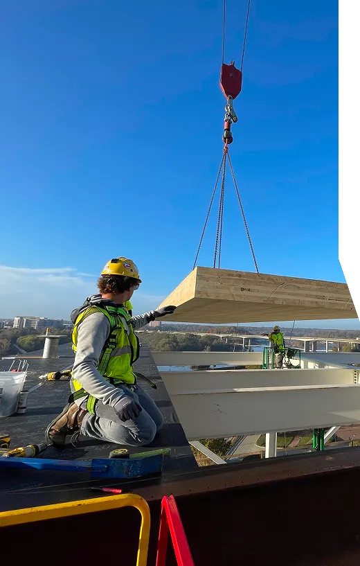 A construction worker guides a large wooden panel being lifted by a crane at a building site.