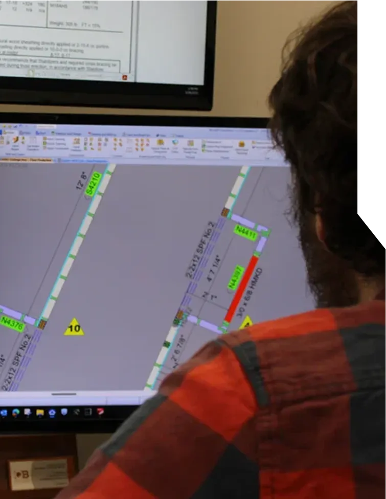 A person in a checkered shirt works at a computer displaying technical diagrams on screen.