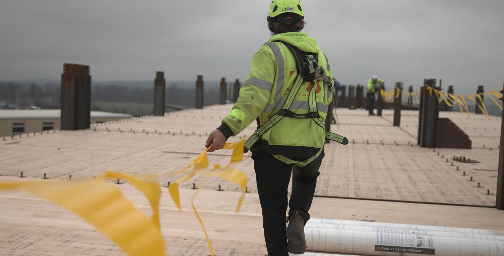 A construction worker in a safety vest is walking across a building site, holding yellow caution tape.