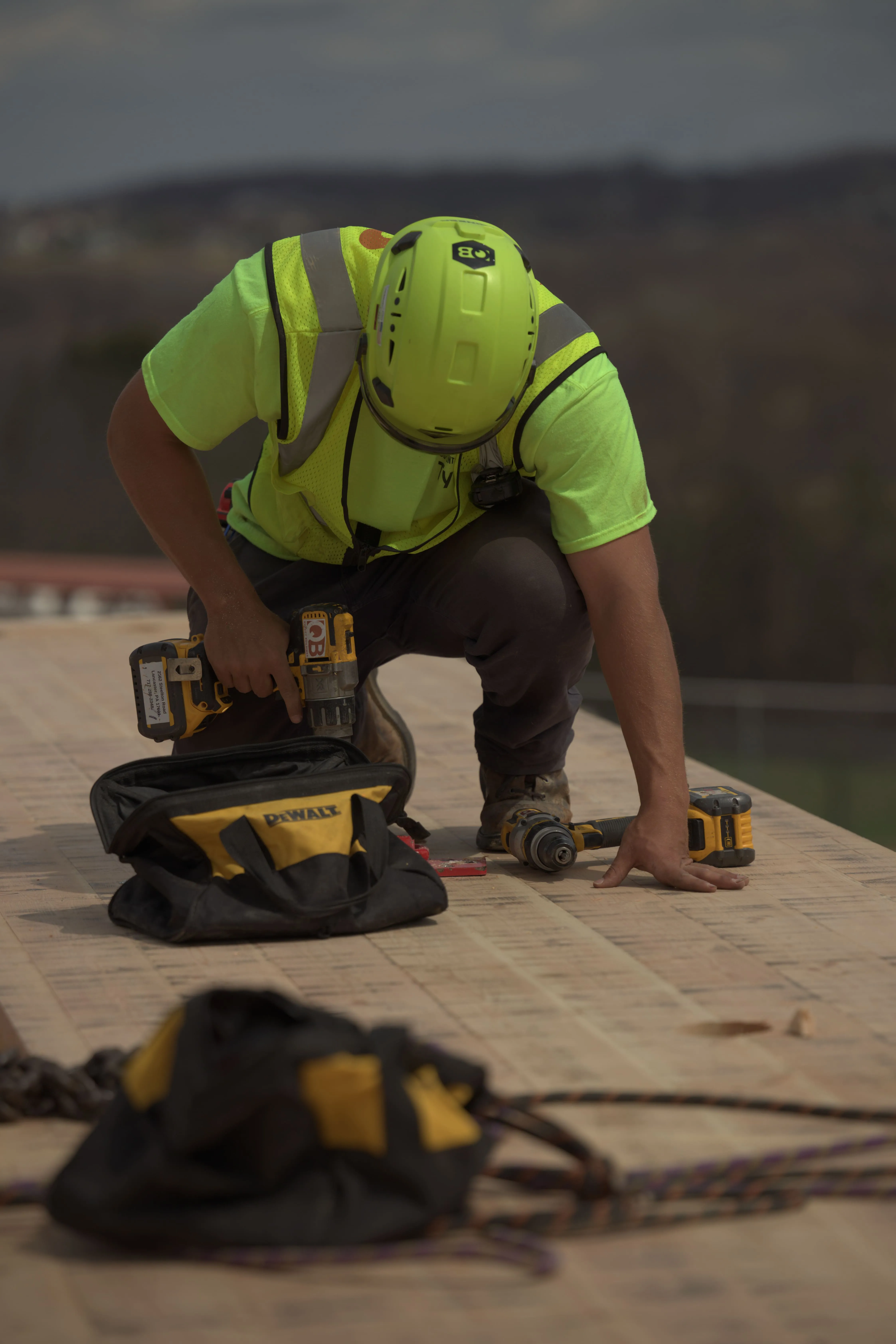 A construction worker in a safety vest and helmet uses a drill on a wooden surface.