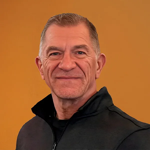 A person with short hair smiling, wearing a dark jacket against a plain orange background.