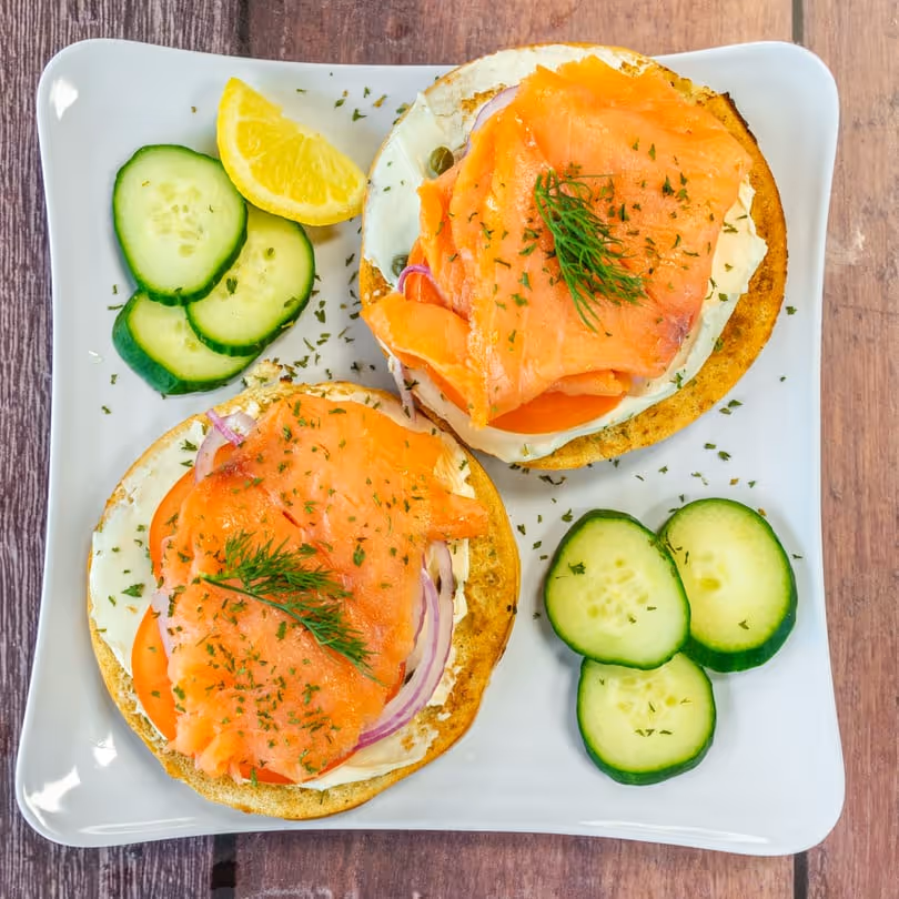 Two smoked salmon bagel halves with cream cheese, tomato, onion, dill, cucumber slices, and lemon wedge on white plate.