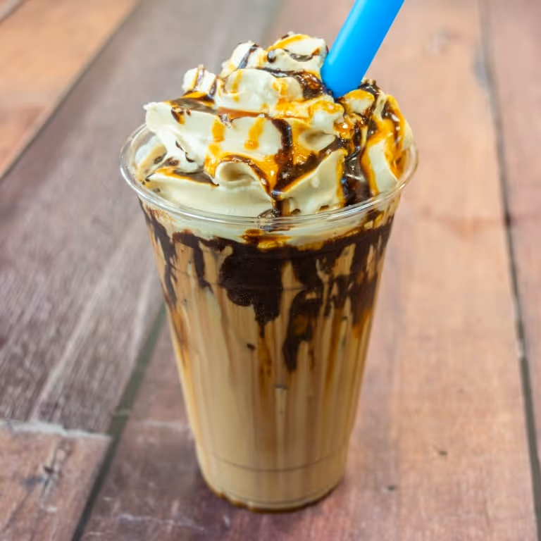 Iced coffee drink in clear cup topped with whipped cream and chocolate and caramel drizzle with blue straw.
