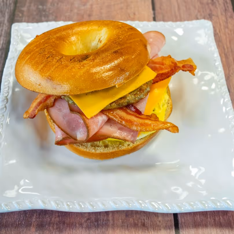Bagel sandwich with bacon, ham, egg, sausage patty, and cheddar cheese on a white plate.