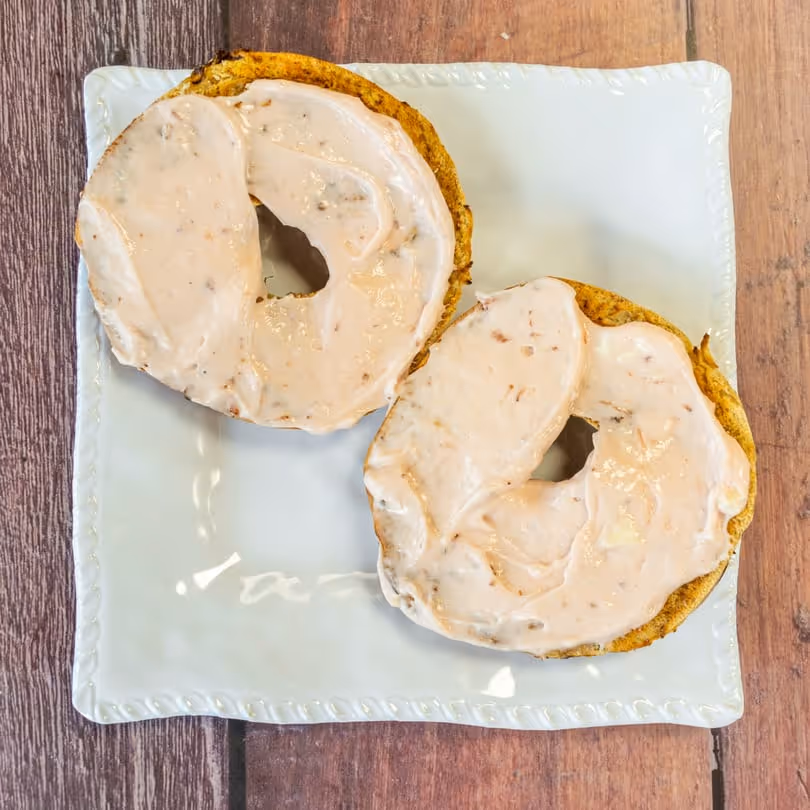 Toasted bagel halves spread with cream cheese on a square white plate over wood surface.